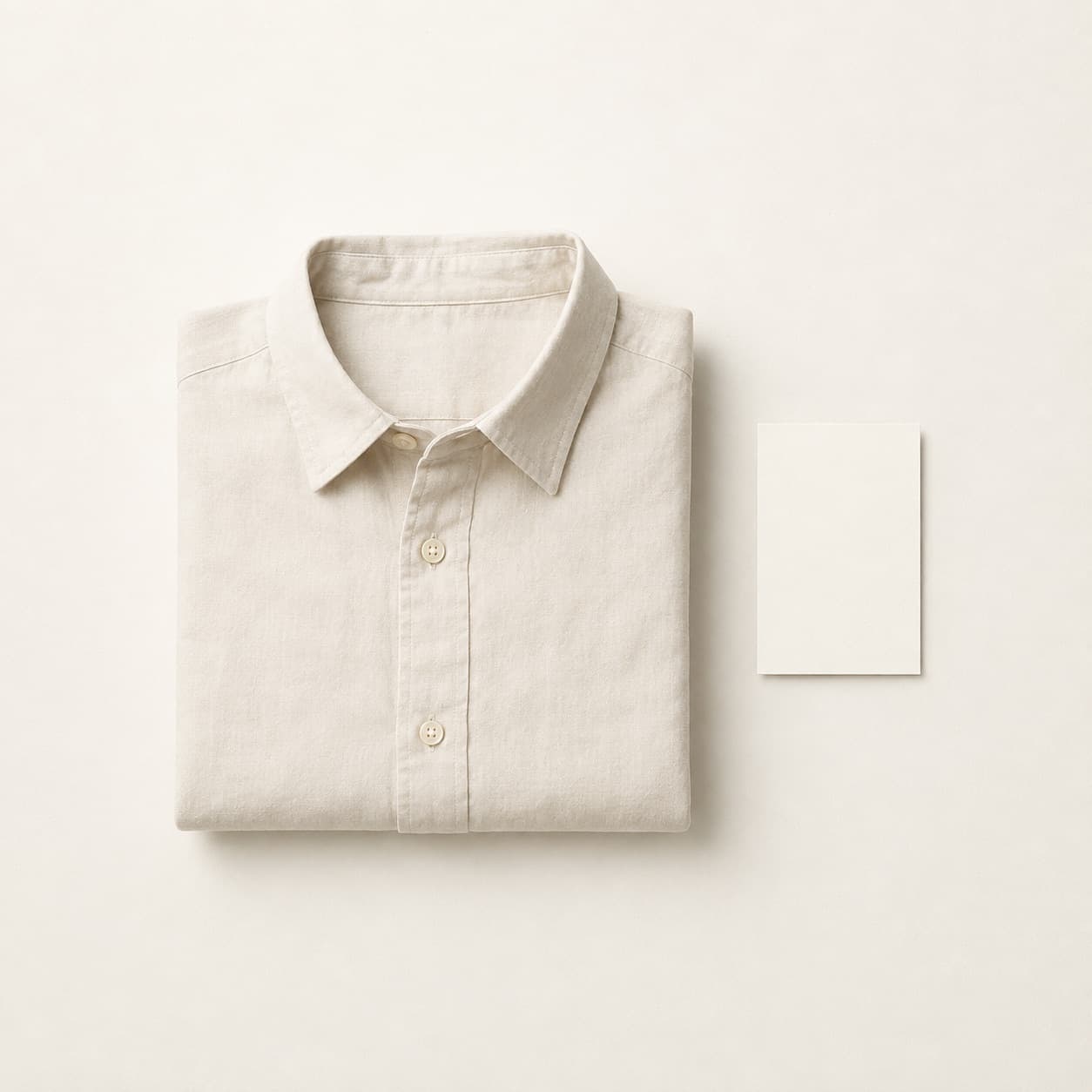 A folded cotton shirt beside a small blank notecard on a pale paper surface